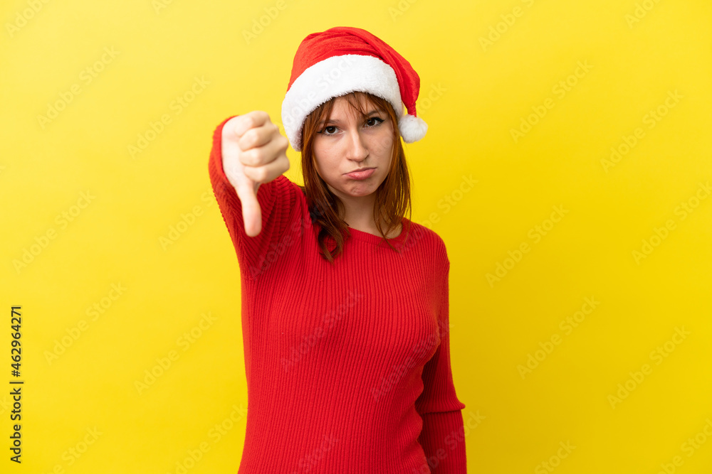 Redhead girl with christmas hat isolated on yellow background showing thumb down with negative expression