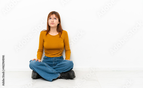 Redhead girl sitting on the floor isolated on white background and looking up