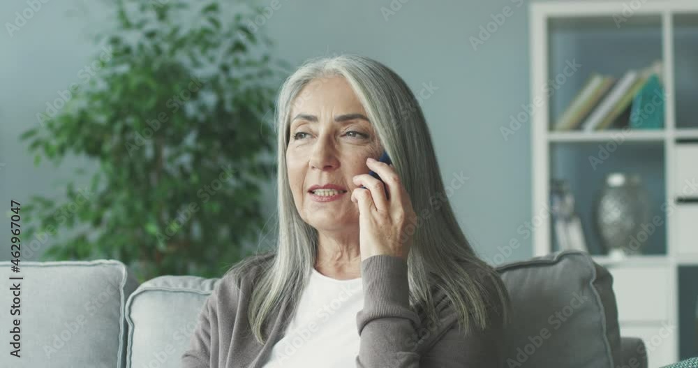 Beautiful gray-haired woman sitting alone at home on couch and talking to friend on mobile phone exchanging news. Beautiful gray-haired woman sitting alone at home on couch and talking to friend on mobile phone exchanging news.