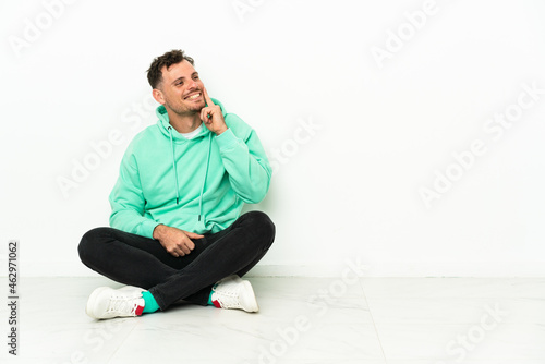Young handsome caucasian man sitting on the floor thinking an idea while looking up