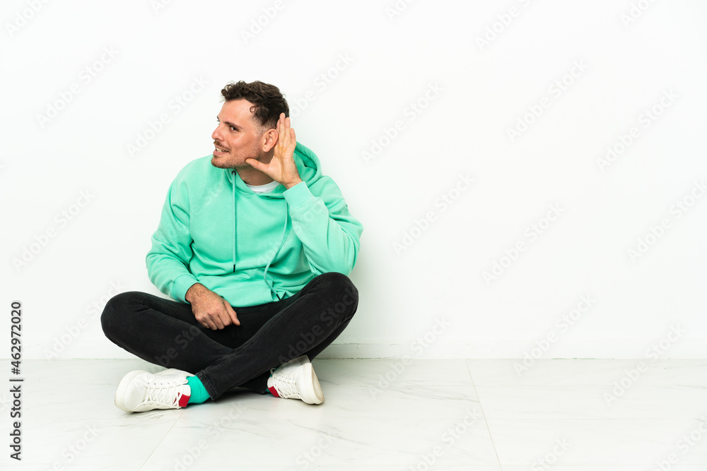 Young handsome caucasian man sitting on the floor listening to something by putting hand on the ear Young handsome caucasian man sitting on the floor listening to something by putting hand on the ear