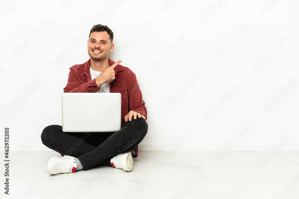 Young handsome caucasian man sit-in on the floor with laptop pointing to the side to present a product Young handsome caucasian man sit-in on the floor with laptop pointing to the side to present a product