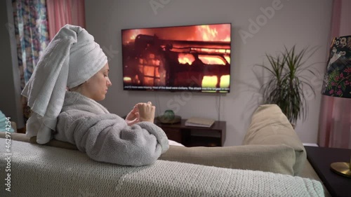 A young Woman is doing a Manicure Sitting on the Sofa in the Living Room with a TV. Presenter Telling Shocking Breaking News with Scenes of Violence, Fires, Protests and Clashes from TV Screen. photo