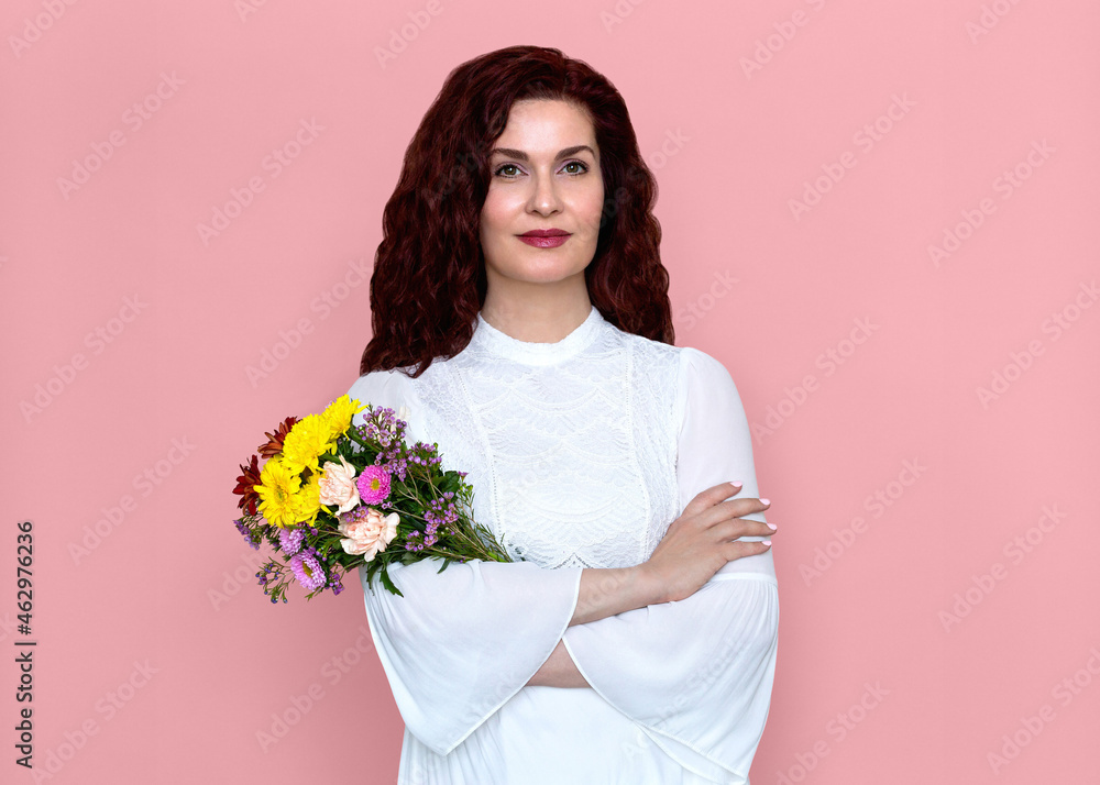 Woman Holding Flowers with Crossed Arms Against Pink Background. Close-up portrait of pretty woman with friendly soft smile in white dress holding bouquet of flowers.