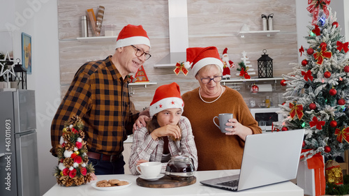 Grandparents with granddaughter greeting remote friends during online videocall meeting celebrating christmas holiday in xmas decorated culinary kitchen. Happy family enjoying winter season together