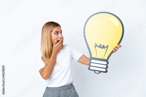 Blonde Uruguayan girl isolated on white background holding a bulb icon with surprised expression