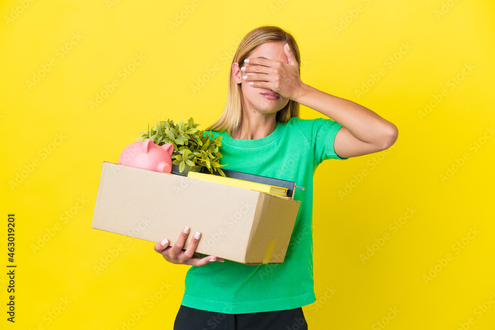 Young Uruguayan girl making a move while picking up a box full of things isolated on yellow background covering eyes by hands. Do not want to see something Young Uruguayan girl making a move while picking up a box full of things isolated on yellow background covering eyes by hands. Do not want to see something