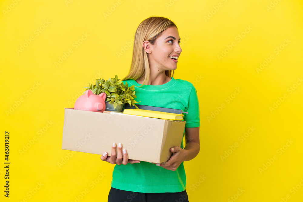 Young Uruguayan girl making a move while picking up a box full of things isolated on yellow background laughing in lateral position Young Uruguayan girl making a move while picking up a box full of things isolated on yellow background laughing in lateral position