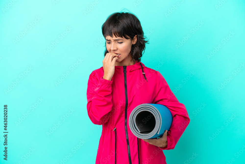 Fototapeta premium Young girl going to yoga classes while holding a mat isolated on blue background having doubts