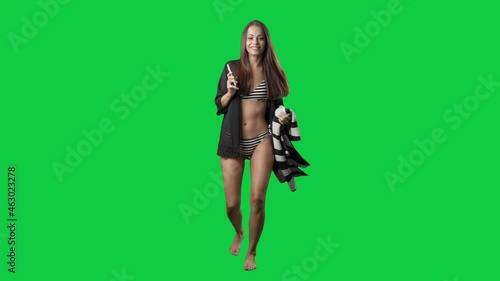 Walking jumpy and dancing young woman in beach wear with towel listening music on phone speaker. Full body on green screen chroma key background