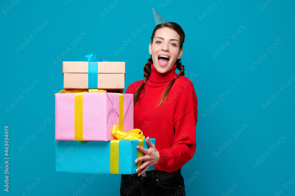 Young woman with party cone on head holding stack of gift and laughing on blue isolated background Young woman with party cone on head holding stack of gift and laughing on blue isolated background