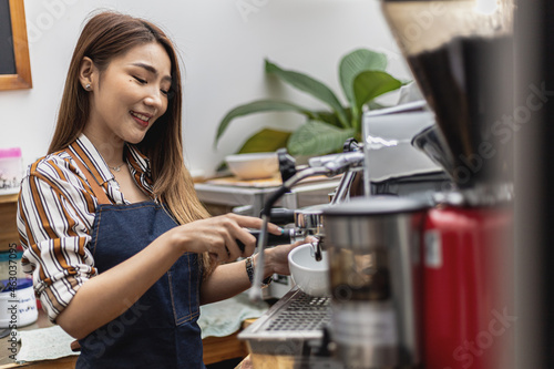 Portrait of a beautiful Asian woman in an apron using a coffee machine, she owns a coffee shop, concept of a food and beverage business. Store management by a business woman.