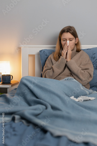 Handsome woman in beige sweater sitting in bed and blows her nose into a paper napkin. Girl suffering from runny nose, rhinitis or sinusitis. Seasonal disease. Treatment at home. Healthcare concept.