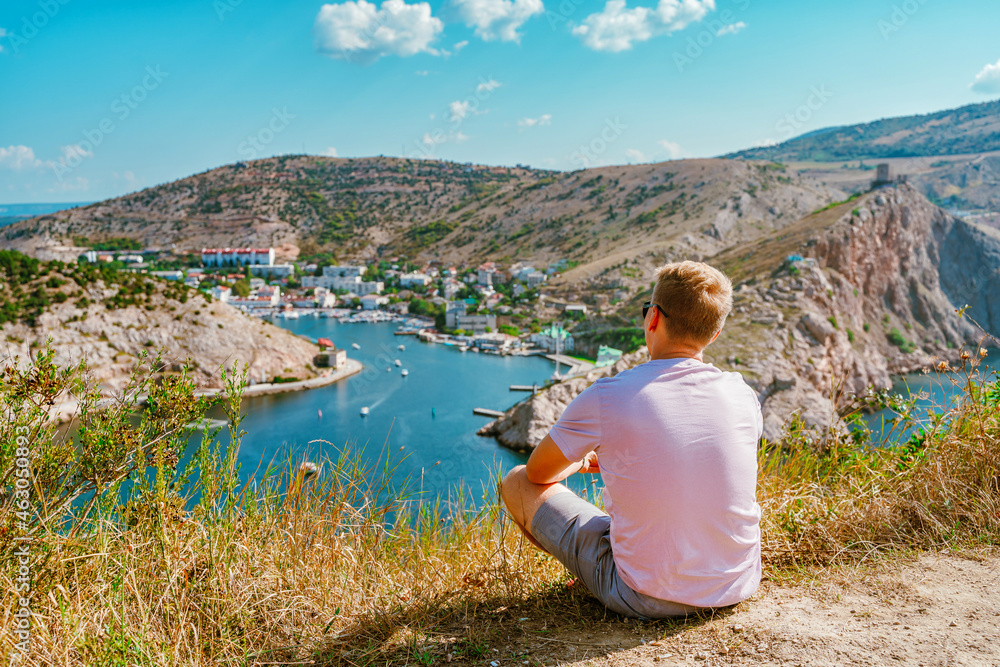 A young blond man enjoys a picturesque landscape with a view of Balaclava with yachts and a colorful bay in summer. Postcard view of the tourist Crimea.