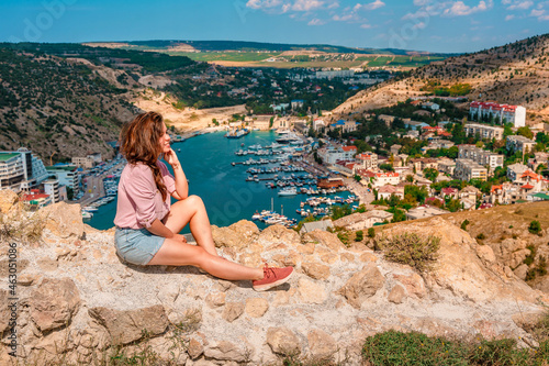 A young beautiful woman admires the picturesque landscape with a view of Balaclava with yachts and a colorful bay in summer. Postcard view of the tourist Crimea.