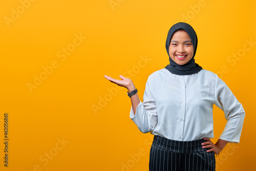 Smiling young Asian woman presenting product and looking camera on yellow background © Sewupari Studio