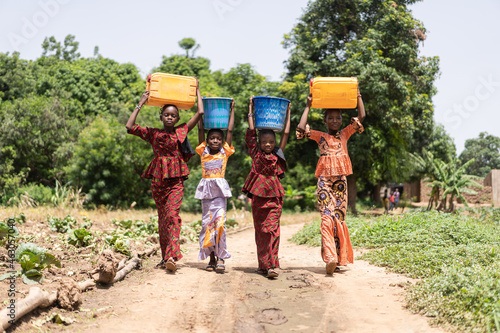 Group of four strong beautifully dressed black African girls carrying water containers on their heads on their way home from the village well