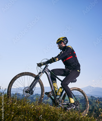 Sportsman in safety helmet and glasses cycling uphill on sunny day with blue sky on background. Bicyclist in cycling suit climbing uphill on mountain bike. Concept of sport and active leisure.