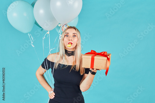 Young woman with gift box and balloons looking at camera and blowing party horn or noisemaker while celebrating birthday on blue background photo