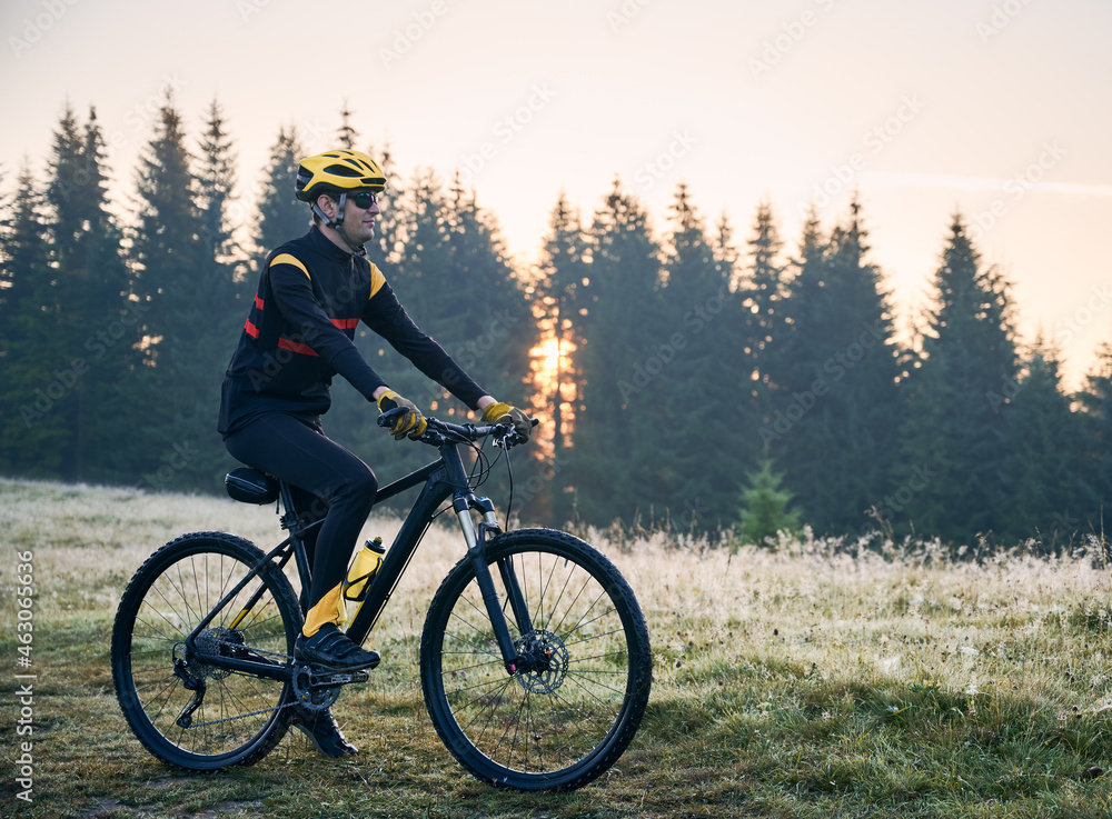 Fototapeta premium Side view of cyclist standing with bike on mountain trail with coniferous trees on background. Man bicyclist enjoying bicycle ride in mountains in the morning. Concept of sport and active leisure.
