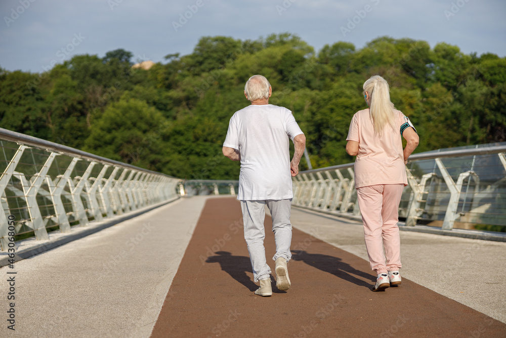 Sportive senior man and woman in tracksuits run together along footbridge Sportive senior man and woman in tracksuits run together along footbridge