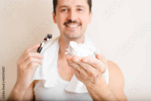 Handsome smiling young man with towel on shoulders holding razor and showing moisturizing shaving foam  close-up. Selective soft focus on white shaving cream. Daily skin care  male beauty concept
