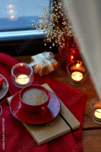 holidays  decoration and celebration concept - cup of coffee in red ceramic cup with saucer  burning candles and christmas gift on window sill at home