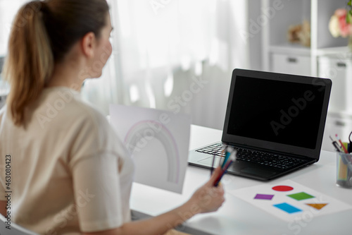 distant education, school and people concept - female elementary school teacher with laptop computer and picture of rainbow having online class at home