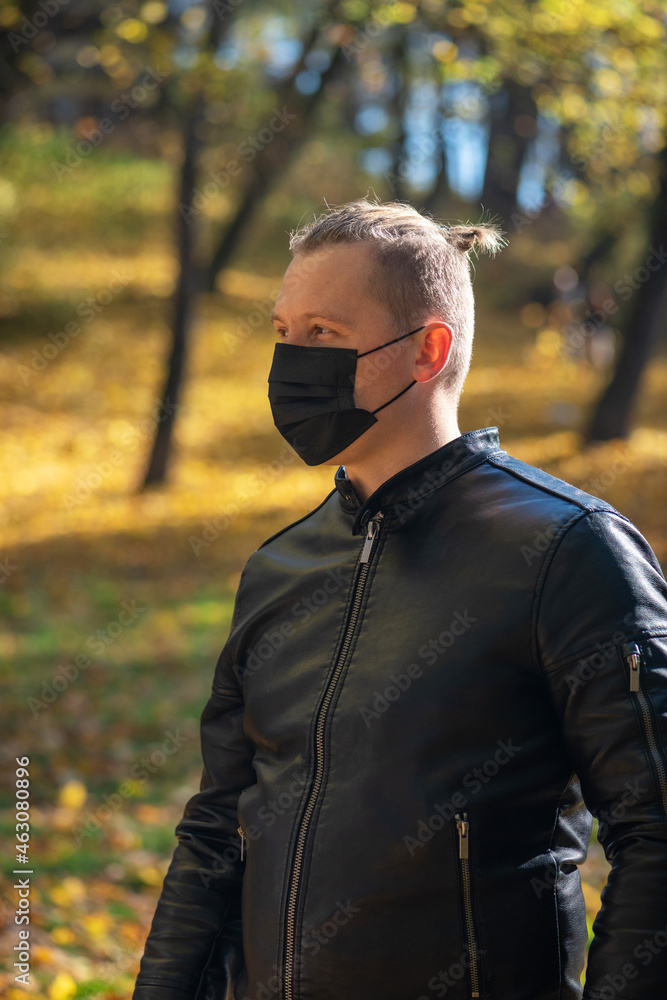 Healthcare, covid-19, coronavirus concept. Man portrait with black leather jacket and top knot hairstyles wearing black face medicine mask in blurry autumn nature background during sunny day Healthcare, covid-19, coronavirus concept. Man portrait with black leather jacket and top knot hairstyles wearing black face medicine mask in blurry autumn nature background during sunny day