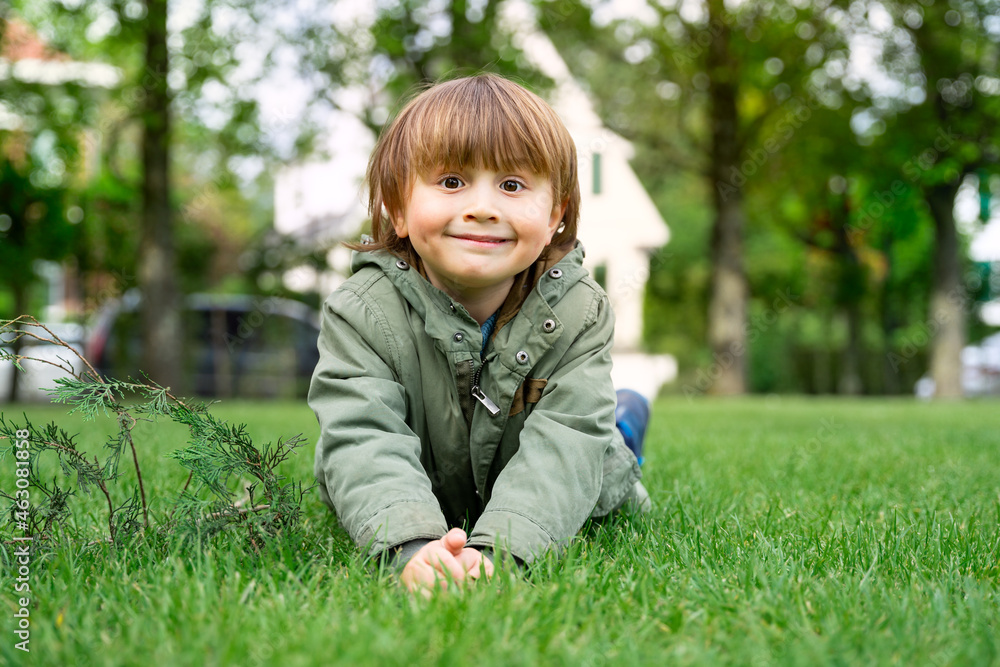Portrait of a little preschool boy wearing green parka lying on the lawn in fresh day. Happy smiling child outdoor. Portrait of a little preschool boy wearing green parka lying on the lawn in fresh day. Happy smiling child outdoor.