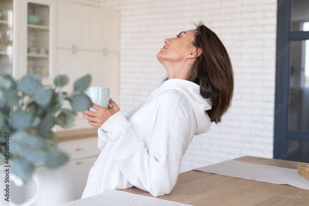 Young woman in a warm white hoodie at home in the kitchen, starts her day with a cup of coffee, posing, in a great mood, smiling Young woman in a warm white hoodie at home in the kitchen, starts her day with a cup of coffee, posing, in a great mood, smiling