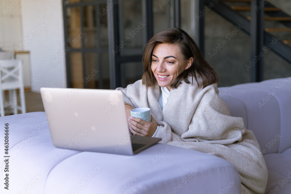Naklejka premium Young woman at home on a sofa in winter under a cozy blanket with a laptop, watching comedy series, laughing, smiling in a winter evening