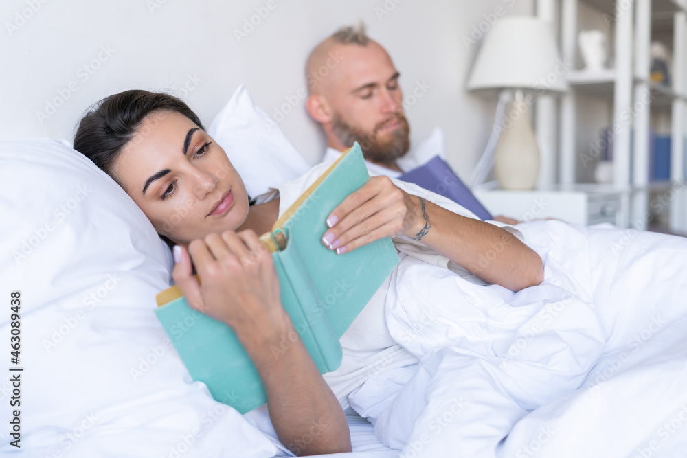 Fototapeta premium Young couple husband with wife at home in bed reading books in autumn winter evening, man fell asleep