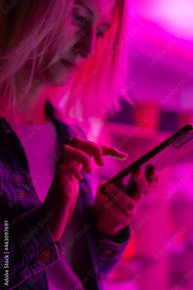 Portrait of a young blonde woman with a mobile phone in her hands in the pink neon light. Pretty young woman in casual street clothes is typing a message on her smartphone. Vertical photo