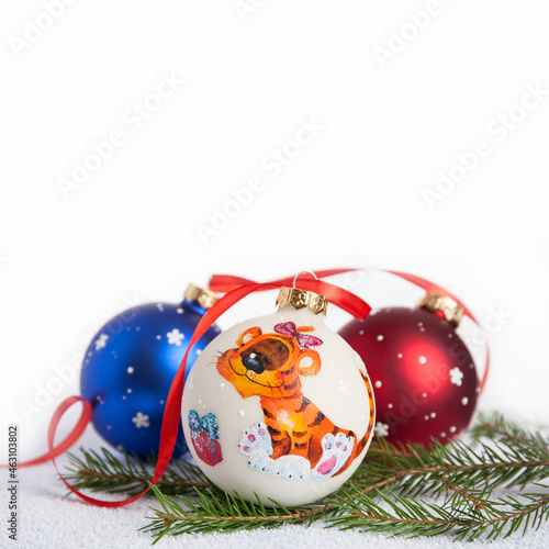 Christmas decoration - hand-painted Christmas balls in white, blue and red colors. One of them is with the symbol of the Year of the Tiger. Blurred background of white color. Fir branches. Red ribbon