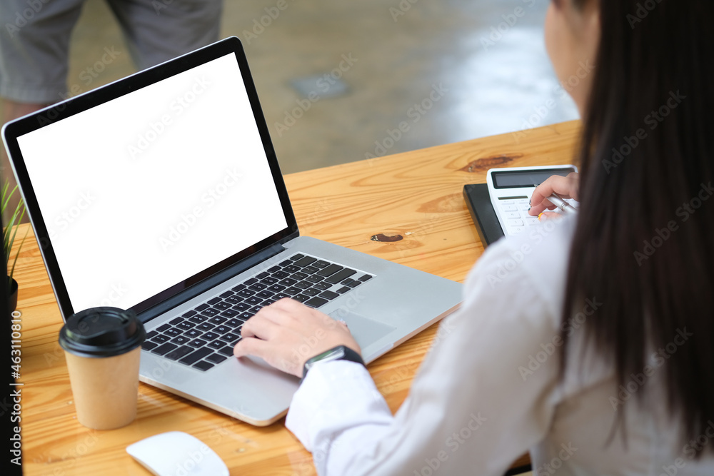 Behind view of a young accountant working with a white screen computer laptop and calculator at the wooden table surrounded by a coffee cup and various equipment. Behind view of a young accountant working with a white screen computer laptop and calculator at the wooden table surrounded by a coffee cup and various equipment.