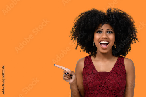 Positive young woman with afro hairdo, points to the side with a cheerful expression, shows something incredible in the blank space, isolated on a orange background