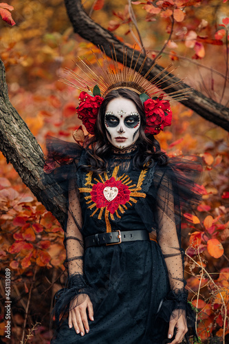 Woman with sugar skull makeup dressed as Santa Muerte is against background of autumn forest.