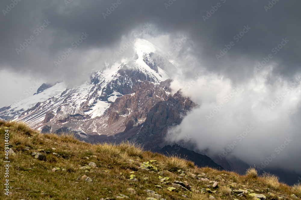 A strong overcast building up above Mount Kazbeg in Caucasus, Georgia. Rainy clouds. There slopes are barren and stony below the snow-capped peak and the Gergeti Glacier. Massive glacier foot