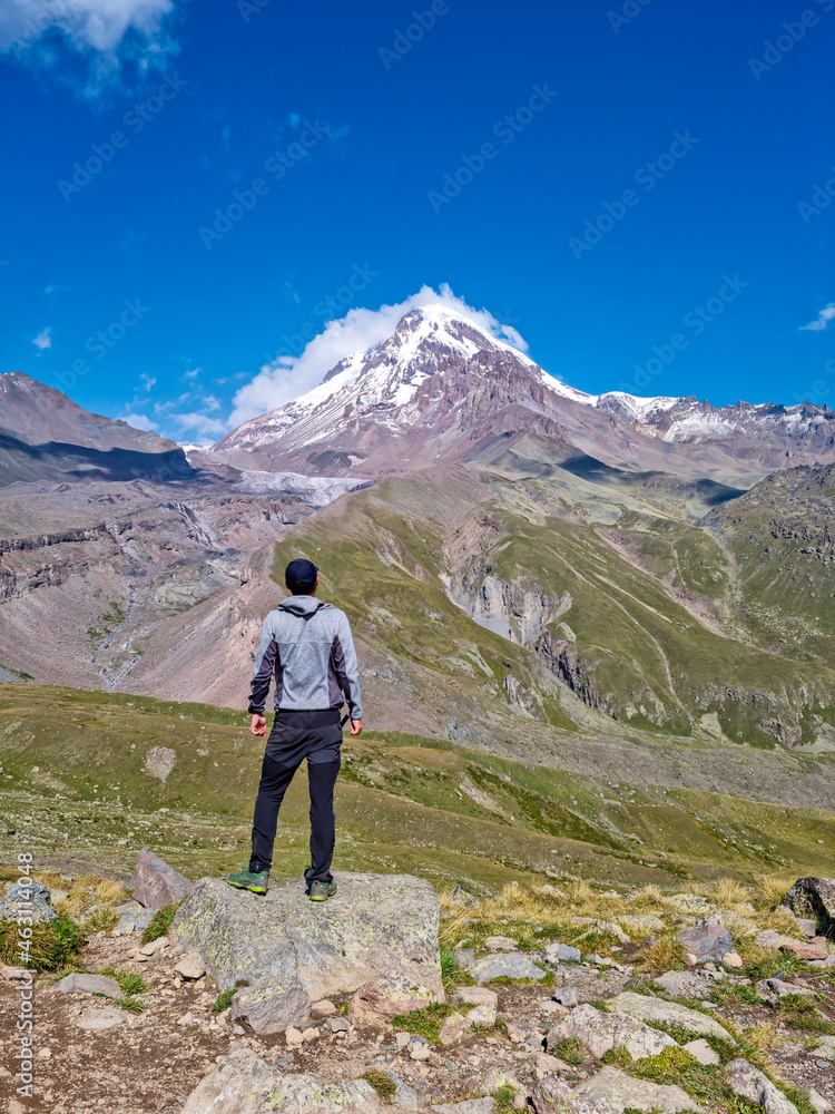 A man enjoying the view on a cloudless view on Mount Kazbeg in Caucasus, Georgia. There are lush pastures and green hills below the snow-capped peak of the glacier. Serenity and Tranquillity. Nature