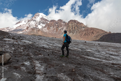A man standing at the foothill of Gergeti Glacier, with the view on Mount Kazbeg, Caucasus, Georgia. Barren slopes below the snow-capped peak. Few clouds gathering around the sharp peak. Tranquillity © Chris