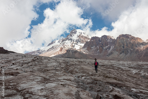 A woman standing at the foothill of Gergeti Glacier, with the view on Mount Kazbeg, Caucasus, Georgia. Barren slopes below the snow-capped peak. Clouds gathering around the sharp peak. Tranquillity