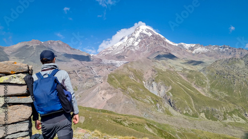 A man leaning agains a stony pillar and enjoying the view on a cloudless view on Mount Kazbeg in Caucasus, Georgia. There are lush pastures and green hills below the snow-capped peak of the glacier.