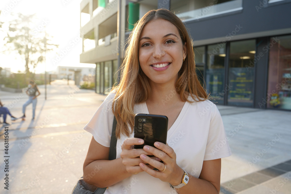 Portrait of cheerful woman looking at camera with phone in city at sunset - obrazy, fototapety, plakaty 