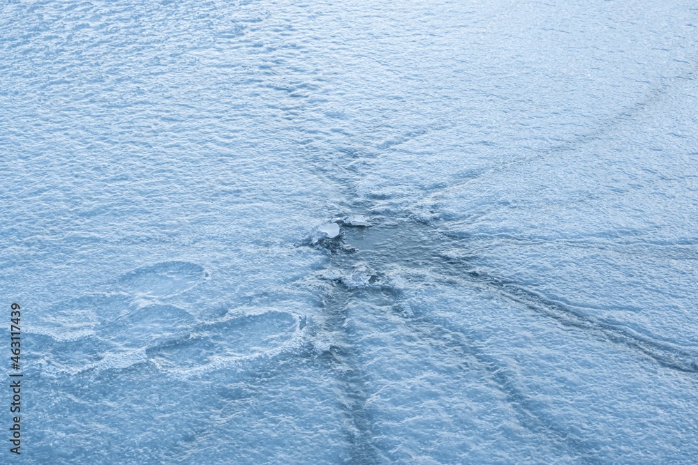 frozen fishing hole with long ice cracks on empty river surface with fisherman footprints, mysterious misty backlight, popular seasonal outdoor hiking detail