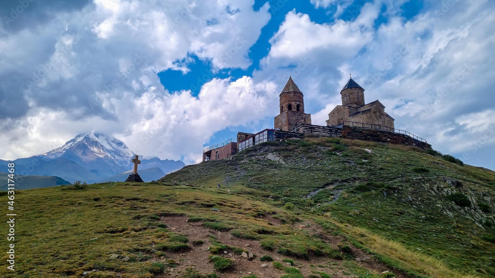 A view on Gergeti Trinity Church in Stepansminda, Georgia. The church is located on a high Caucasian mountain. Clear and blue sky above the church. In the back there are high mountain chains. A view on Gergeti Trinity Church in Stepansminda, Georgia. The church is located on a high Caucasian mountain. Clear and blue sky above the church. In the back there are high mountain chains.