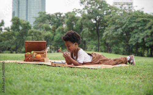 Little African kid boy crying and holding a sore finger when playing xylophone toys alone with sad face at park and lying down on mat with picnic basket on weekend