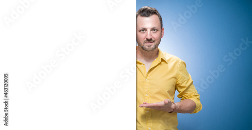 Happy young women holding white board over blue background, dresses in yellow shirt. Handsome guy showing blank empty paper billboard with blank space for text