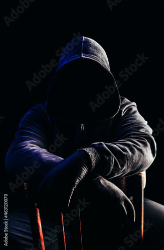 Photo of scary horror stranger stalker man in black hood and clothing sitting on chair on dark background.