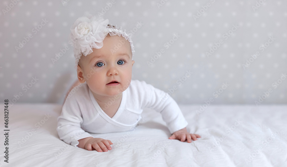 Toddler baby girl in a white bodysuit lying on bed at home looking at the camera. Copy space for text, mock up, banner Toddler baby girl in a white bodysuit lying on bed at home looking at the camera. Copy space for text, mock up, banner